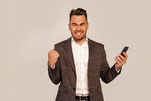 excited man with mobile phone showing win gesture isolated on grey