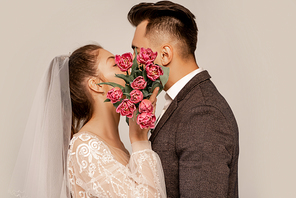 young newlyweds kissing while obscuring faces with tulips isolated on grey