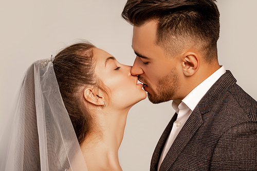 side view of young newlyweds kissing with closed eyes isolated on grey