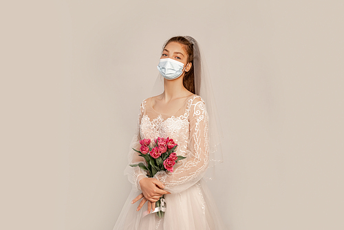 young bride in safety mask holding fresh tulips isolated on grey with lilac shade