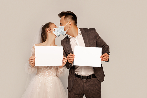 newlyweds in safety masks holding blank cards while kissing isolated on grey