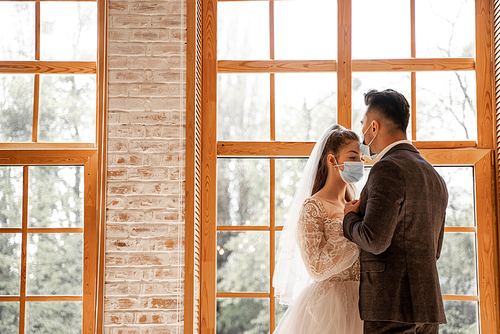 newlyweds in protective masks near large windows at home
