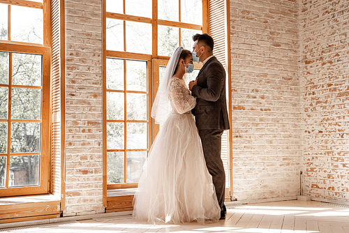 full length view of young newlyweds in protective masks standing near large windows