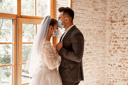 newlyweds in medical masks standing with closed eyes near large window