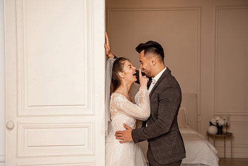 side view of laughing bride touching nose of happy groom near entrance to bedroom