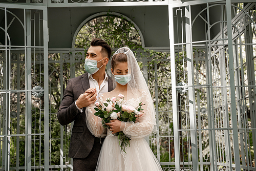 young man in medical mask holding hand of bride with wedding bouquet in park