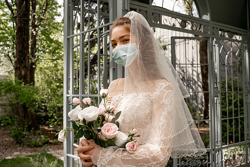 elegant bride in protective mask  while holding wedding bouquet outdoors