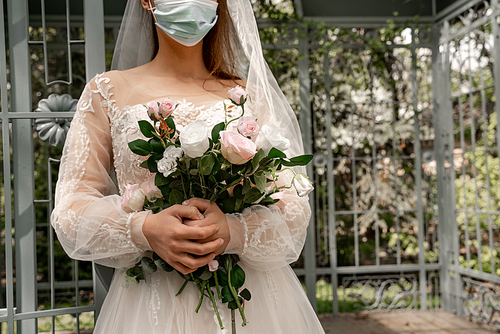 cropped view of elegant bride in medical mask holding wedding bouquet in park