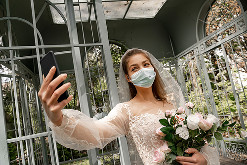 woman in wedding dress and medical mask taking selfie on smartphone in park