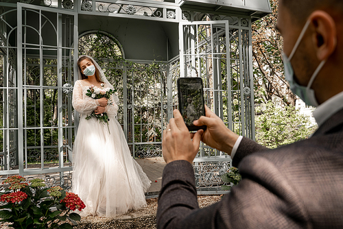 blurred man in medical mask taking picture of bride near alcove in park