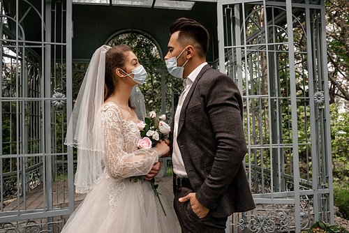side view of young bride and groom in medical masks looking at each other outdoors