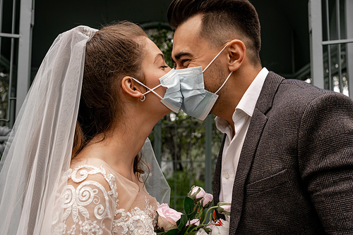side view of elegant newlyweds in medical masks kissing in park