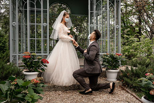 man in medical mask standing on knee near elegant bride in park