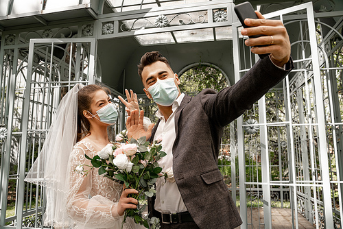 newlyweds in medical masks taking selfie on mobile phone in park