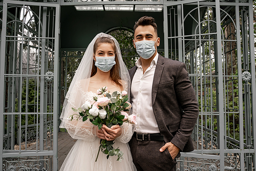 elegant newlyweds in medical masks  near alcove in park