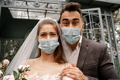 excited newlyweds in safety masks with bride and groom lettering  in park