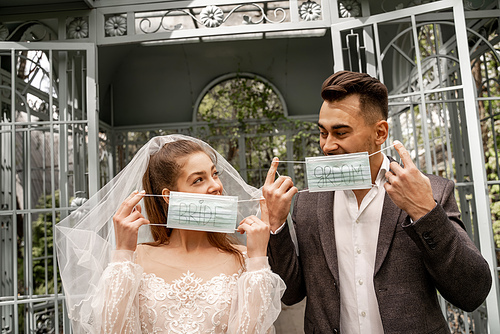 young couple looking at each other while holding medical masks with bride and groom lettering
