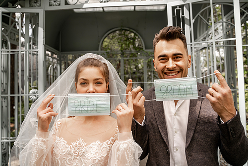 happy man and woman holding medical masks with bride and groom lettering