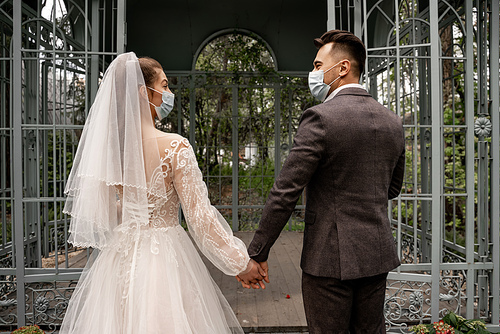 back view of bride and groom in medical masks holding hands near alcove in park