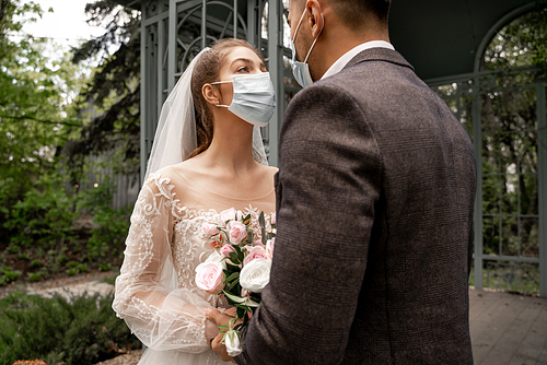 bride in medical mask looking at groom while holding bouquet outdoors