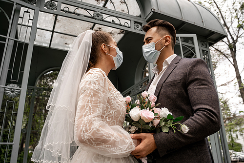 low angle view of newlyweds in protective masks looking at each other outdoors