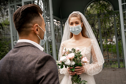 young bride in medical mask holding wedding bouquet near groom on blurred foreground