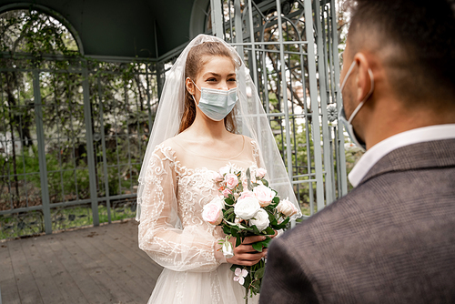 elegant bride in medical mask holding wedding bouquet near blurred groom in park