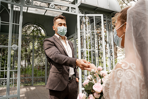 young newlyweds in safety masks holding hands in park on blurred foreground