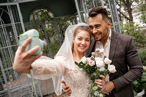 cheerful bride taking selfie with groom on digital camera in park, blurred foreground