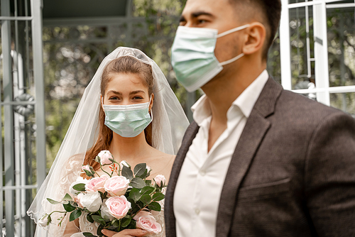 young woman with wedding bouquet  near blurred groom in medical mask