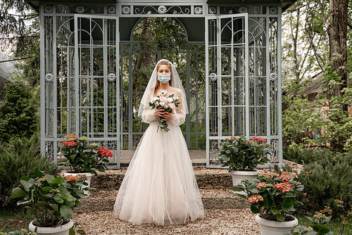 full length view of elegant bride in safety mask holding wedding bouquet near alcove in park