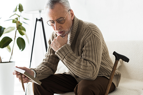 pensive retired man holding photo frame while sitting on sofa near walking stick