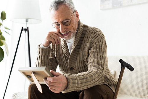 cheerful retired man holding photo frame while sitting on sofa near walking stick