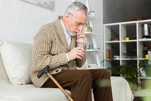 senior man taking pill while sitting on sofa near walking cane