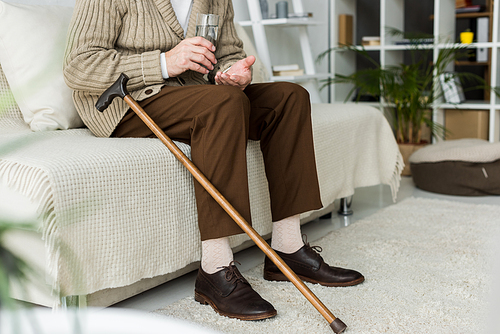 cropped view of retired man holding pill while sitting on sofa near walking cane