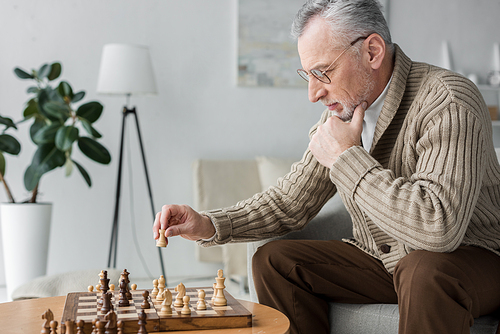 retired man in glasses thinking while playing chess at home