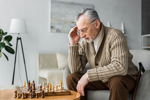 pensive retired man in glasses thinking while playing chess at home