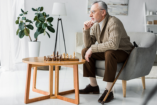 senior man in glasses thinking while sitting near chess board at home