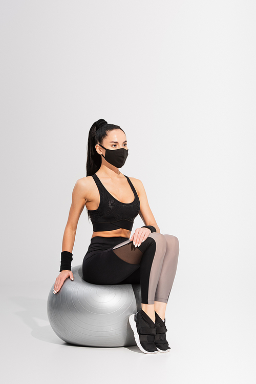 young sportswoman in black protective mask sitting on fitness ball on grey