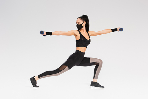 full length of young sportswoman in black protective mask exercising with dumbbells on grey