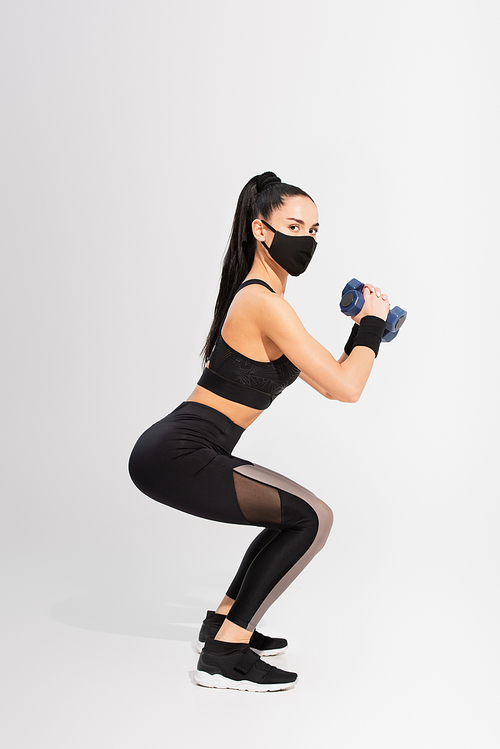 full length of sportswoman in black protective mask exercising with dumbbells on grey