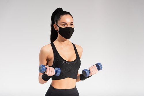 sportswoman in black protective mask exercising with dumbbells on grey