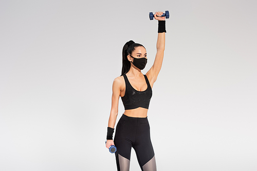sportswoman in black protective mask working out with dumbbells on grey