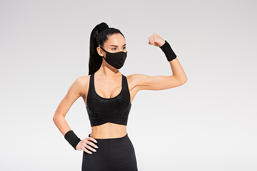 young woman in sportswear and black protective mask showing muscle on grey