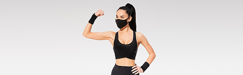young woman in sportswear and black protective mask showing muscle isolated on grey, banner