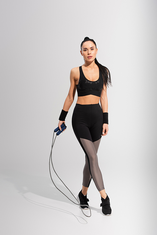 full length of young woman in sportswear holding skipping rope on grey