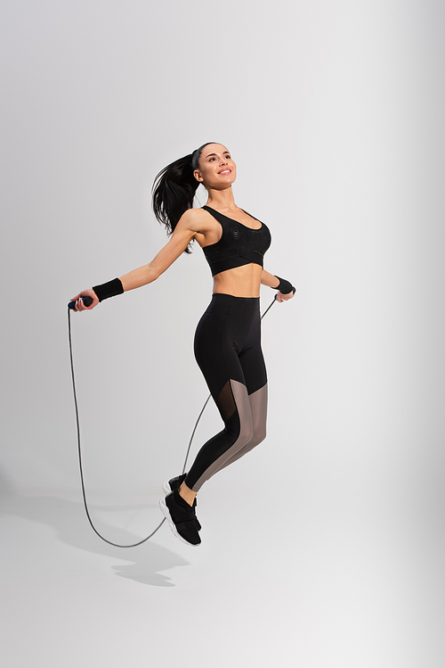 full length of happy sportswoman exercising with skipping rope on grey
