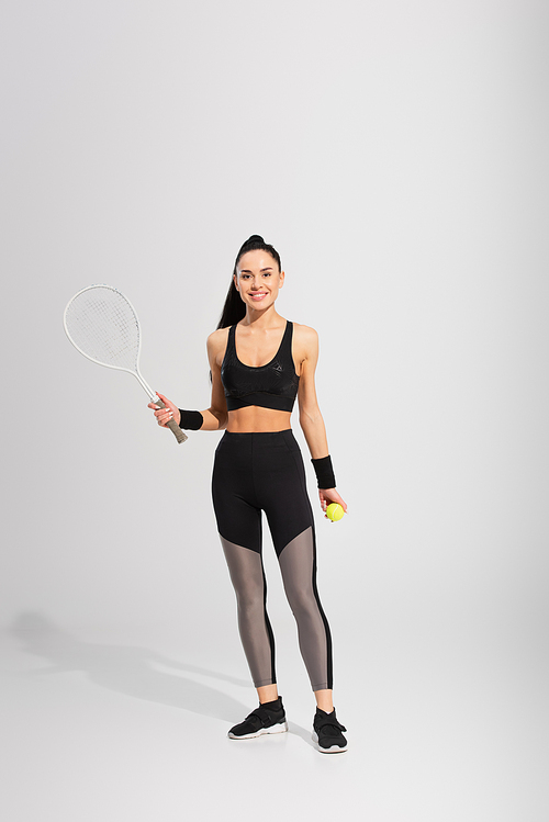 full length of joyful sportswoman holding tennis racket and ball on grey