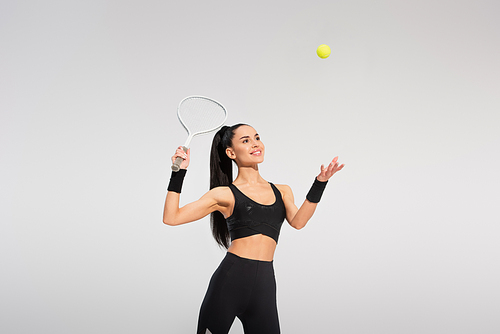 happy sportswoman holding tennis racket and throwing ball while playing on grey