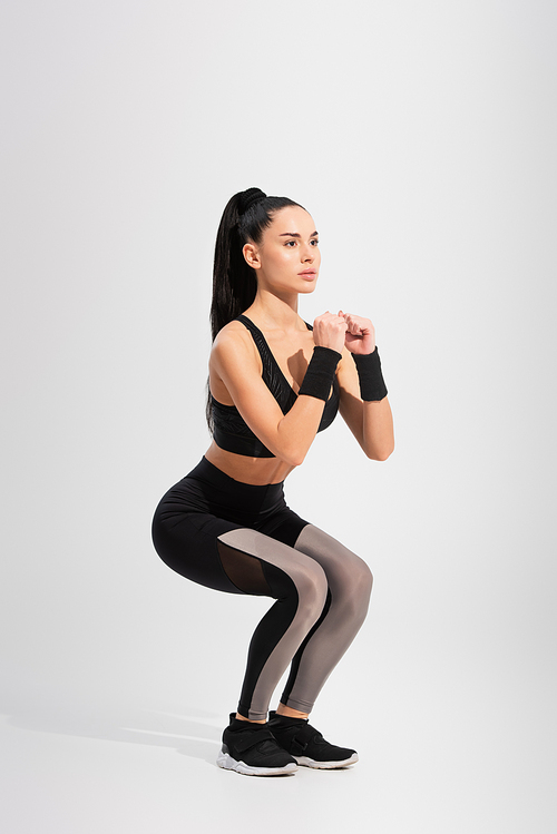 full length of young brunette sportswoman doing squat exercise on grey